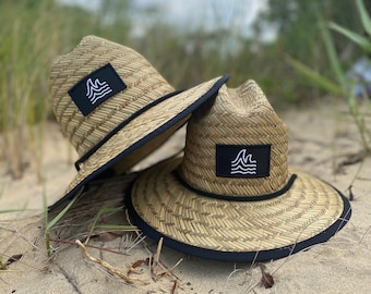May include: Two straw hats with a black brim and a black square patch featuring a white wave design. The hats are resting on sand with some grass in the background. The hats are a light tan color.