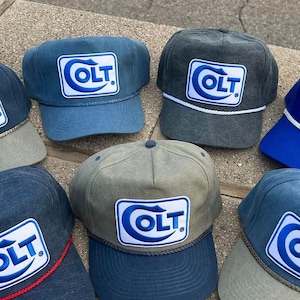 Peut inclure: Plusieurs casquettes de baseball bleues, grises et vertes. Chaque casquette arbore le logo "COLT" dans un rectangle blanc avec un contour bleu. Certaines casquettes ont un détail de corde autour du bord.