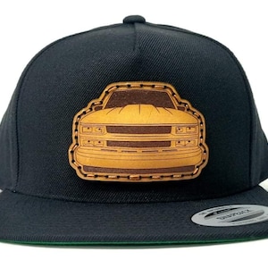 May include: Black and brown baseball cap with a leather patch of a vintage truck on the front. The patch is stitched around the edges and has a brown leather background with a gold truck.