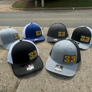 May include: Six trucker caps in shades of grey, black and blue. Each cap has a patch with the number "33" and a cross design. The caps have a mesh back and a curved peak.