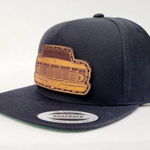 Puede incluir: Gorra de béisbol negra con un parche de cuero marrón que presenta un diseño de parrilla de camión vintage. La gorra tiene una parte inferior verde y una etiqueta blanca que dice "The Authentic Snapback Yupoong Since 1974".