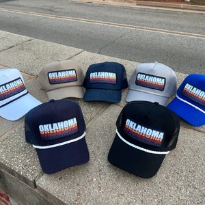 May include: Seven trucker hats with a mesh back and a colorful "Oklahoma" patch on the front. The hats are in various colors, including white, beige, blue, gray, black, and blue.