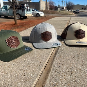 Puede incluir: Tres gorras de camionero con un parche de cuero marrón en la parte delantera. Las gorras son de diferentes colores: verde, gris y beige.