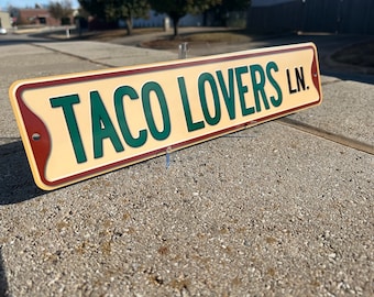 May include: A rustic-style metal sign with a cream background, brown border, and green text that reads "Taco Lovers Ln."