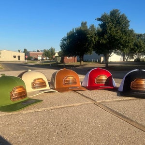 Könnte beinhalten: Fünf verschiedene farbige Trucker-Caps mit einem braunen Leder-Patch eines Autos auf der Vorderseite. Die Caps sind in einer Reihe auf einer Betonfläche angeordnet.