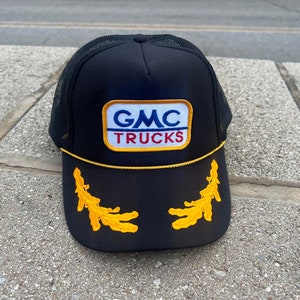 May include: Black trucker hat with a yellow and white GMC Trucks patch and yellow embroidered wheat stalks on the front.