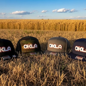Op de afbeelding: Vier baseballpetten in zwart, camouflage, grijs en marineblauw, elk voorzien van een witte patch met de rood omrande tekst OKLA. De petten hebben een gestructureerde kroon en een platte klep, met een kleine sticker op elke klep zichtbaar.