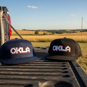 Puede incluir: Dos gorras snapback, una azul marino y otra negra, cada una con la palabra OKLA bordada en blanco con un borde rojo en el panel frontal. Ambas gorras tienen una corona estructurada y una visera plana, colocadas una al lado de la otra.