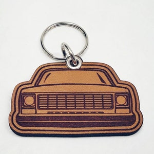 Puede incluir: Un llavero de cuero marrón con una silueta detallada de frente de una camioneta clásica. La camioneta tiene una parrilla y faros negros.