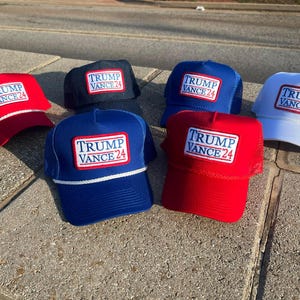 Może przedstawiać: Sześć niebieskich, czerwonych i białych czapek trucker z białym naszywką z napisem "TRUMP VANCE 24" na każdej czapce.