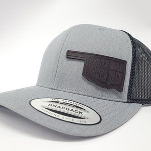 May include: A gray and black trucker hat with a brown leather patch that says "Sooner State Sooner Pride" and an outline of the state of Oklahoma. The hat has a black mesh back and a gray front with a "The Authentic Snapback Yupoong Since 1974" label.