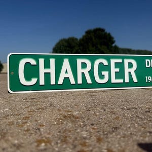 Puede incluir: Un letrero de calle verde y blanco que dice "CHARGER DR 1966". El letrero rectangular tiene letras blancas y un borde blanco. Está montado en una pared de hormigón, con un cielo azul y árboles al fondo.