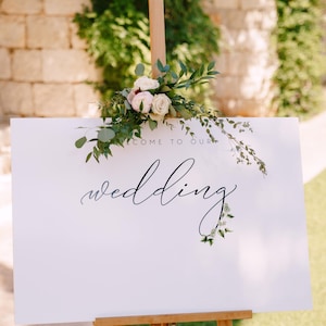 May include: White wedding sign with black calligraphy that reads "Welcome to our wedding" with greenery and white flowers on top. The sign is on a wooden easel.