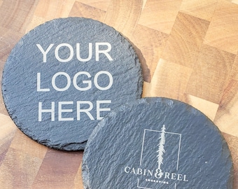 May include: Two round, dark gray slate coasters. One coaster has the text "YOUR LOGO HERE" in white. The other coaster has a logo with a tree and the text "CABIN & REEL ENGRAVING" and "WWW.CABINANDREEL.COM" in white.