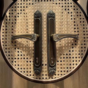 Puede incluir: Dos manillas de puerta de estilo antiguo con detalles ornamentados y acabado en latón. Las manillas están montadas en un asiento de silla de mimbre tejido.
