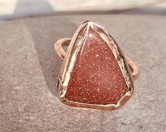 May include: A gold-toned ring with a triangular, reddish-brown stone. The stone has a glittery, speckled appearance. The ring's band is a simple, rounded design, and the stone is set within a gold-colored frame.
