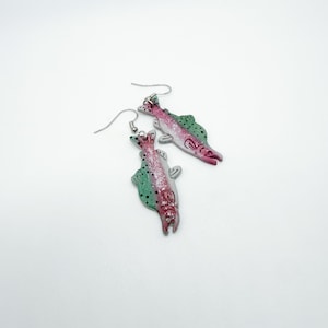 May include: A pair of fish-shaped earrings. Each earring features a silver hook and a colorful fish design with pink, green, and silver accents. The fish have a detailed scale pattern and are set against a white background.