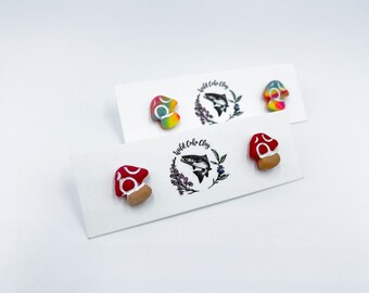 May include: A set of three mushroom-shaped earrings. Two earrings are rainbow colored with white dots and one earring is red with white dots. The earrings are displayed on a white card with the text "Wild Coho Clay".