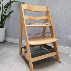 Könnte beinhalten: Ein helles Holzhochstuhl mit Sitz, Fußstütze und Sicherheitsgurt. Es hat eine Metallstange und eine grüne Netzkiste. Der Stuhl ist für Säuglinge und Kleinkinder konzipiert und hat eine natürliche Holzoberfläche.