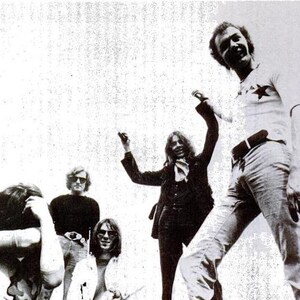May include: A black and white photo of a band with five members. The band members are all wearing casual clothing and are smiling. The photo is taken from a slightly low angle, and the band members are looking up at the camera.