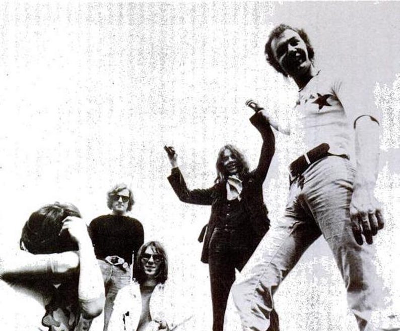 May include: A black and white photo of a band with five members. The band members are all wearing casual clothing and are smiling. The photo is taken from a slightly low angle, and the band members are looking up at the camera.