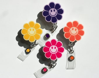 May include: A collection of flower-shaped badge reels in various colors: yellow, purple, orange, and pink. Each reel features a smiling face and a clear plastic clip for attaching to clothing. The reels are designed for holding ID badges.