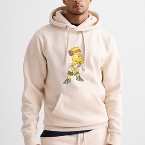 May include: A cream-colored hoodie with a cartoon image of Bart Simpson wearing a bandana and holding a slingshot. The hoodie has a drawstring hood and a kangaroo pocket.