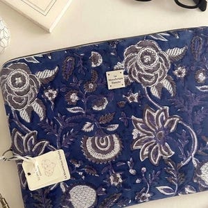 Peut inclure: Une housse d'ordinateur rectangulaire bleue avec un motif floral blanc et gris. Une petite étiquette métallique carrée est centrée sur le devant avec le texte La Manufacture Blanche. Une étiquette en papier blanc avec un cœur géométrique est attachée.