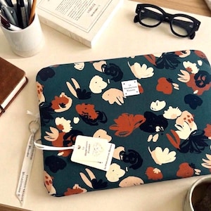 Peut inclure: Une housse d'ordinateur rectangulaire avec un fond bleu sarcelle foncé orné d'un motif floral abstrait dans des tons orange rouille, crème et noir. Une petite étiquette carrée blanche indique La Manufacture Blanche, avec une étiquette attachée.
