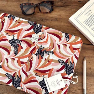 Peut inclure: Une housse d'ordinateur rectangulaire avec un motif répétitif de toucans noirs parmi des feuilles stylisées orange, roses et blanches. La housse comporte une petite étiquette carrée blanche avec du texte et une étiquette blanche sur la tirette.