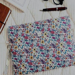 Peut inclure: Une housse d'ordinateur portable rectangulaire matelassée avec un motif floral vibrant composé de fleurs sauvages roses, jaunes, bleues et vertes sur fond blanc. Elle comprend une petite étiquette de marque carrée et une tirette en tissu blanc.