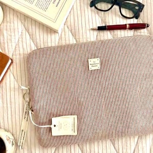Peut inclure: Une housse d'ordinateur rectangulaire avec un motif à fines rayures rouges et blanches. Une petite étiquette en tissu blanc est cousue au centre avec le texte "La Manufacture Blanche". Une étiquette blanche assortie est fixée à la tirette.