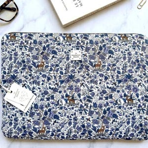 Peut inclure: Une housse d'ordinateur rectangulaire avec un motif dense de fleurs et de forêt dans des tons de bleu, blanc et marron. Le tissu présente de petites figures de cerfs parmi les arbres et les fleurs. Une étiquette blanche indique La Manufacture Blanche.
