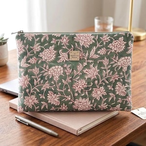 Könnte beinhalten: Eine grüne Laptoptasche mit einem floralen Muster in Rosa und Weiß. Das Design zeigt Blumen und Blätter. Ein kleines Holzschild ist angebracht. Die Tasche liegt auf einem Notizbuch und einem Holztisch.