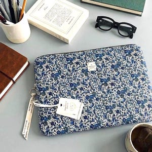 Peut inclure: Une housse d'ordinateur portable rectangulaire avec un motif floral dense dans des tons de bleu, blanc et noir. Elle comprend une petite étiquette de marque blanche, une fermeture éclair avec une tirette en tissu et une étiquette rectangulaire blanche.
