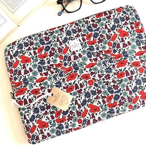 Könnte beinhalten: Eine rechteckige Laptoptasche mit einem dichten Blumenmuster aus roten Mohnblumen, dunkelblauen Blüten und hellgrünen Blumen auf weißem Grund. Ein kleines weißes Stoffetikett trägt die Aufschrift La Manufacture Chasse. Ein beiges Etikett hängt daran.