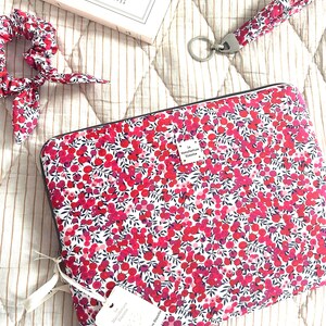 Può includere: Una custodia rettangolare per laptop, un elastico per capelli abbinato e un cinturino da polso, tutti caratterizzati da un vivace motivo floreale di bacche rosse e rosa con foglie verde scuro su sfondo bianco. È attaccata un'etichetta bianca con il testo La Manufacture Blanche.