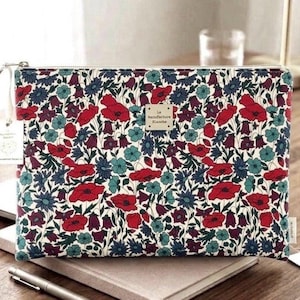 Puede incluir: Un estuche rectangular con cremallera que presenta un denso estampado floral de amapolas rojas, flores verde azulado y flores de color púrpura oscuro sobre un fondo crema. Una pequeña etiqueta rectangular en el centro dice La Manufacture Blanche.