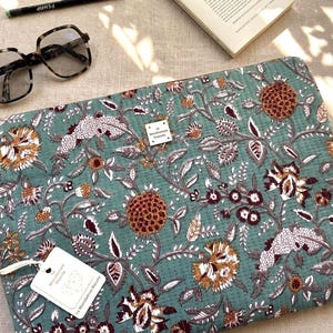 Könnte beinhalten: Eine rechteckige, gesteppte Laptoptasche aus salbeigrünem Stoff mit einem dichten Blumenmuster in Rost-, Braun- und Weißtönen. Ein kleines, quadratisches weißes Etikett mit dem Text La Manufacture Bisou ist mittig auf der Vorderseite angebracht.