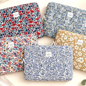 May include: Five rectangular laptop sleeves featuring various floral patterns in red, blue, yellow, and white. Each sleeve has a white zip pull and a small rectangular white label with black text that reads LA MANUFACTURE BLOSSOM.