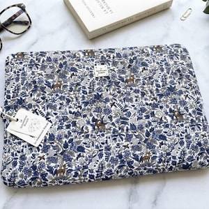 Könnte beinhalten: Eine rechteckige Laptoptasche mit einem dichten Blumen- und Waldtiermuster in Blau-, Weiß- und Brauntönen. Ein kleines weißes rechteckiges Etikett mit dem Text La Manufacture Bleue ist an der Vorderseite angebracht, zusammen mit einem weißen Papieranhänger.