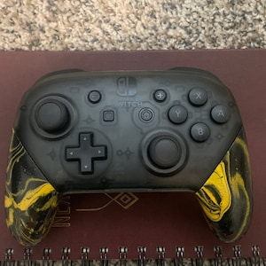 May include: A black Nintendo Switch controller with yellow and black marbled grips. The controller has a black and white design on the front.
