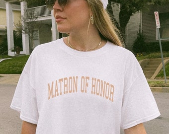 May include: A white t-shirt with the text "MATRON OF HONOR" printed in peach on the front. The person wearing the shirt is also wearing a white baseball cap and sunglasses.