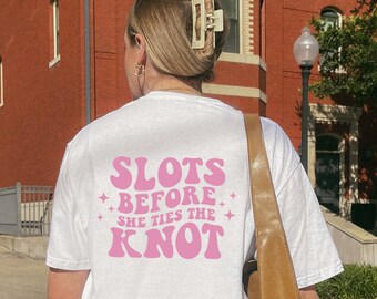 May include: White t-shirt with pink text that reads "Slots Before She Ties The Knot" in a retro font.