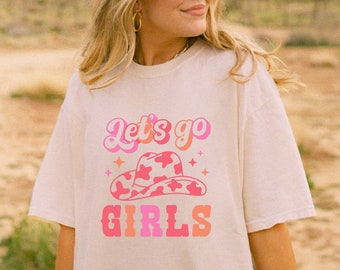 May include: A white t-shirt with a pink and orange graphic design that says "Let's go girls" with a pink and white cowgirl hat with white stars.