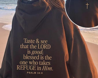 May include: A dark brown hoodie with the text "Taste & see that the LORD is good; blessed is the one who takes REFUGE in Him. PSALM 34:8" printed on the back. The person is wearing a tan baseball cap and a gold cross necklace.