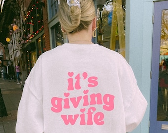 May include: A white sweatshirt with the text "it's giving wife" in pink letters with hearts. The person wearing the sweatshirt is walking down a brick sidewalk.