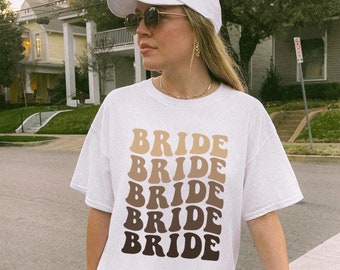 May include: A white t-shirt with the word "BRIDE" repeated five times in a gradient of brown and beige. The text is in a retro font style.