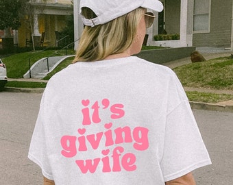 May include: White t-shirt with the phrase "it's giving wife" in pink lettering. The shirt is a casual style, and the text is centered on the back. A white baseball cap is also visible.