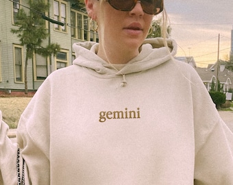 May include: A beige oversized hoodie with a brown embroidered "gemini" on the front. The hoodie has a large pocket on the front and a black chain strap.